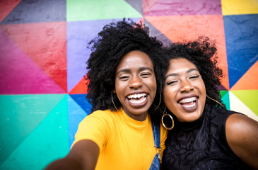 Two women hug each other and laugh while facing the viewer, in front of a colorful background