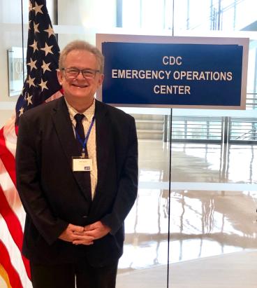 Mark Schleiss standing in front of a U.S. flag and a sign that says, "CDC Emergency Operations Center"